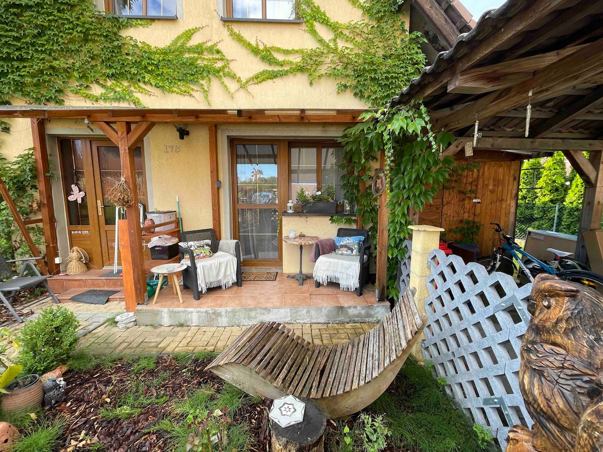 An inviting entrance features a covered terrace adorned with simple seating arrangements and decorative elements. Surrounding greenery enhances the atmosphere, while a wooden sculpture and garden path draw attention to the home's welcoming facade. Bicycles and storage spaces are accessible nearby.