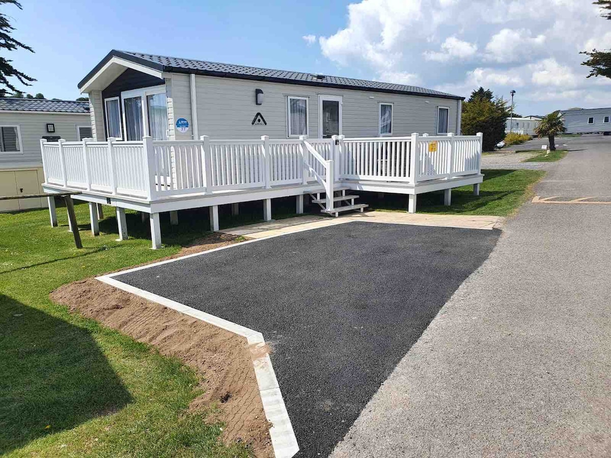 A newly constructed caravan is set on a grassy area, featuring a spacious wooden deck with white railings. Steps lead to the entrance, while a newly paved area offers additional parking space. The sky is partly cloudy, adding to the bright and open setting.