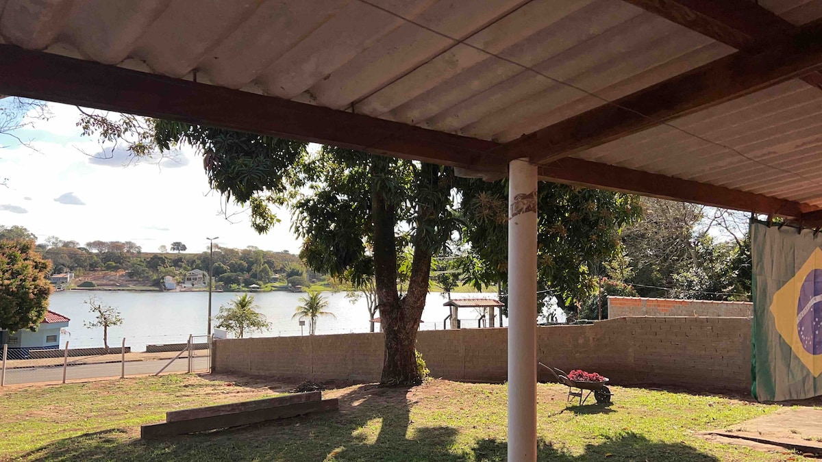 A view from a shaded porch reveals a tranquil water scene, framed by large trees. The calm surface of the water reflects the surrounding landscape, with a distant structure visible along the shoreline. The space offers a relaxing connection to nature.