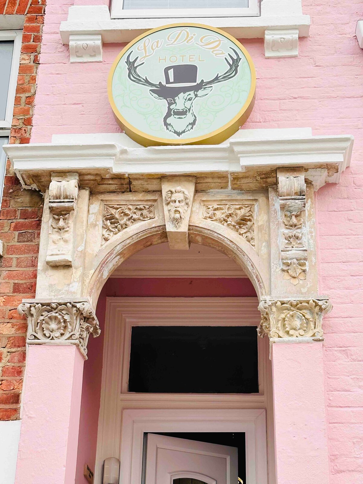 The hotel entrance features an ornate archway made of intricate stone carvings, contrasting beautifully with the pastel pink facade. Above, a circular sign displays the hotel's name alongside a stylized deer wearing a top hat, adding a unique touch to the entrance.