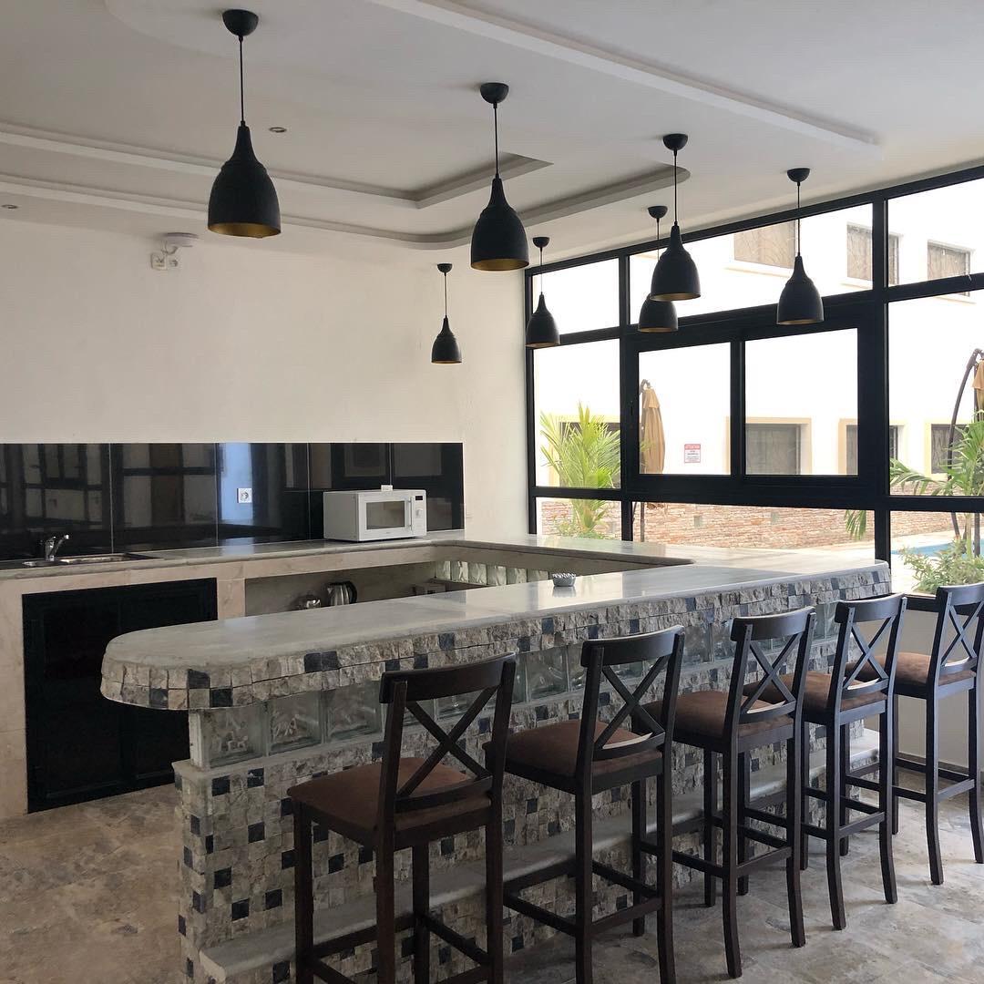 A modern kitchen is presented with a spacious island bar featuring a unique stone finish. Six wooden bar stools are positioned around the island. Large windows allow natural light to fill the space, while overhead pendant lights add a contemporary touch.