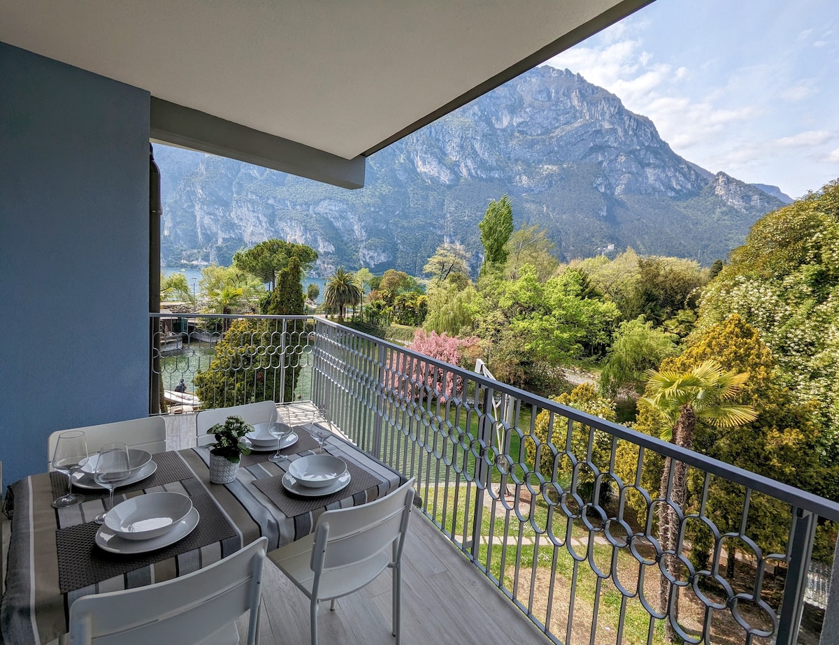 A balcony offers outdoor dining with a table set for four. Lush greenery surrounds the area, complemented by distant mountain views and a serene lake. The space is brightened by natural light, highlighting the balcony's modern railing design.