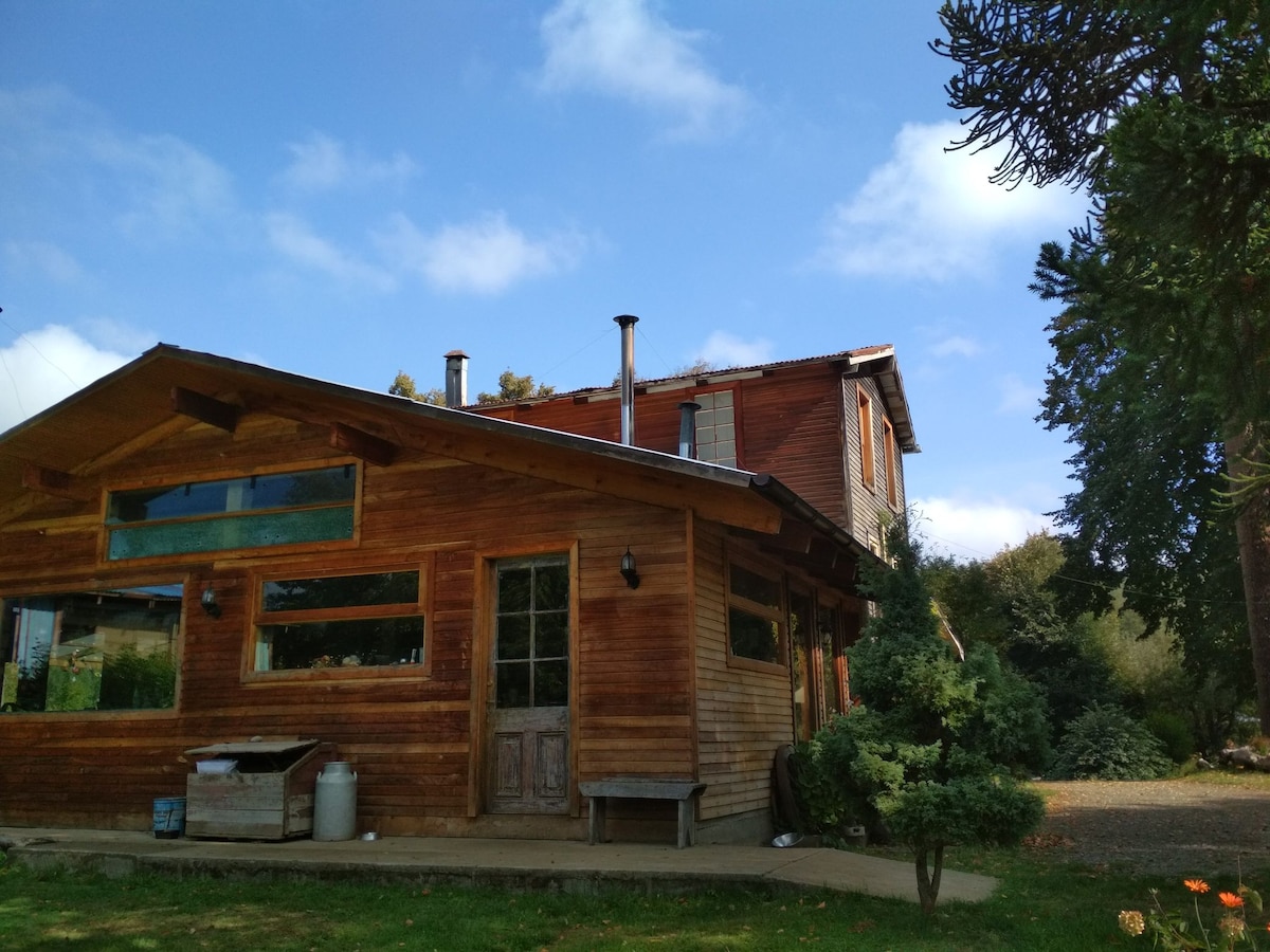 The exterior of a wooden house is showcased, featuring a combination of textured wood and large windows. A chimney is visible, and the surrounding landscape includes greenery and well-maintained shrubs. The sky above is partly cloudy, contributing to a serene outdoor environment.
