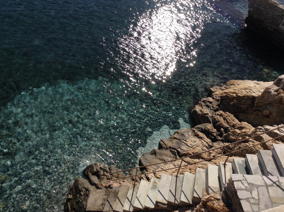 A view of the crystal-clear Aegean Sea reveals smooth, rocky shores and gently undulating water. Sunlight reflects off the surface, creating a shimmering effect. A stone stairway, leading down towards the seaside, is visible on the right side of the image.