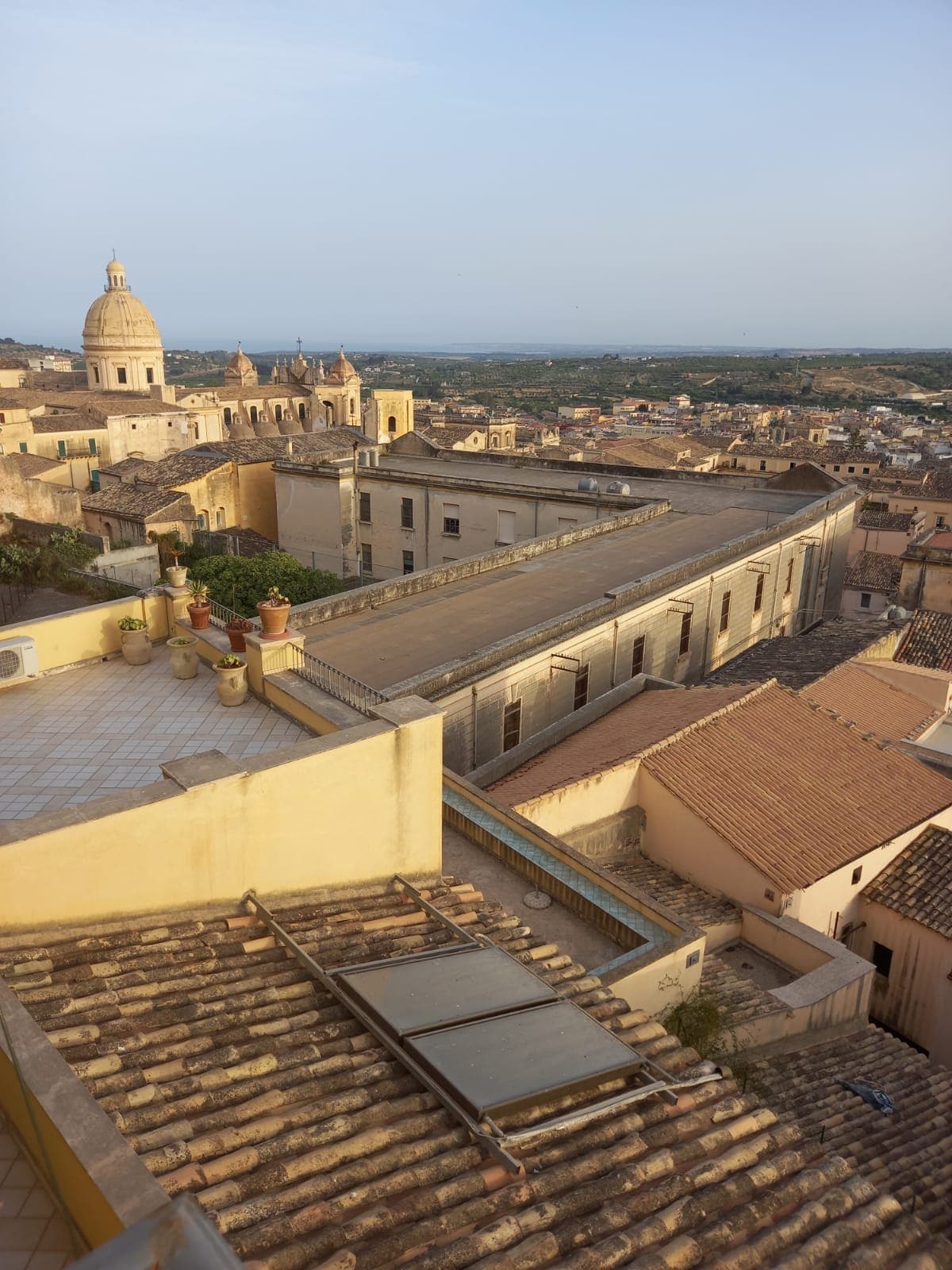 A panoramic view captures the rooftops of historic buildings, showcasing terracotta tiles and varying architectural styles. In the distance, a prominent dome is visible against the sky, while rolling hills extend beyond the cityscape.