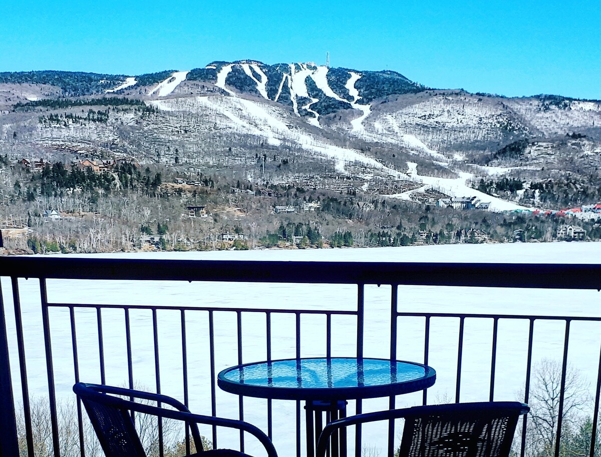 A balcony is shown featuring a small round table and two chairs, overlooking a frozen lake and the snowy slopes of Mont-Tremblant in the background. The bright blue sky enhances the scenic winter view.