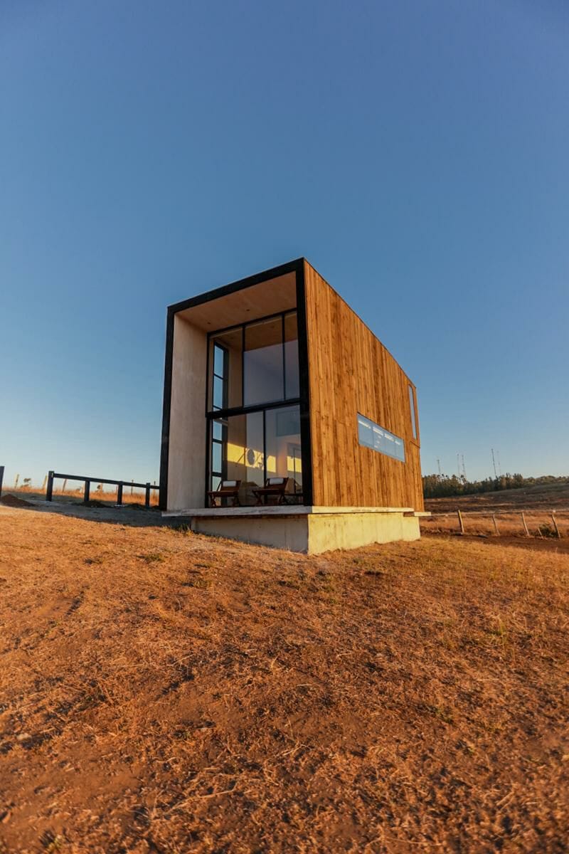 An architectural structure is presented with a combination of wood and concrete materials. Large glass windows afford views of the surroundings, while a wooden deck is visible, adding to the outdoor space. The setting includes a dry landscape under a clear blue sky.