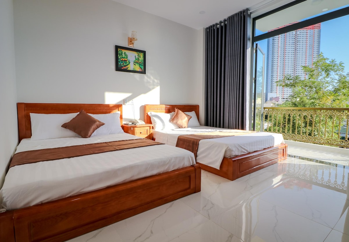 A modern hotel room features two spacious beds with neutral linens, complemented by decorative brown throw pillows. Natural light fills the room through large sliding doors, which open to a balcony overlooking greenery. A small bedside table and a framed artwork enhance the inviting space.