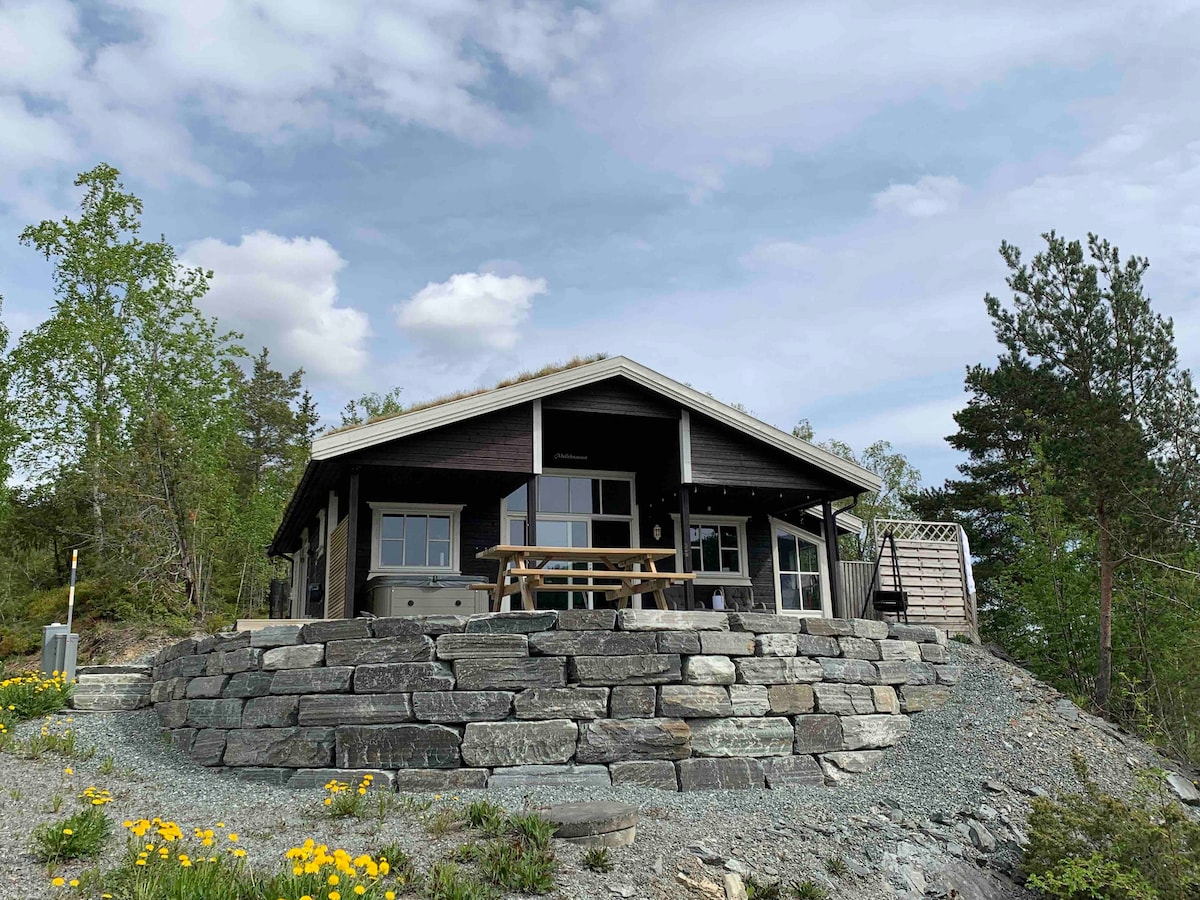 A charming cabin is situated on a stone foundation, surrounded by greenery. Large windows allow for natural light, and a wooden picnic table is placed on the spacious outdoor terrace. A peaceful landscape is visible, providing a sense of tranquility and comfort.