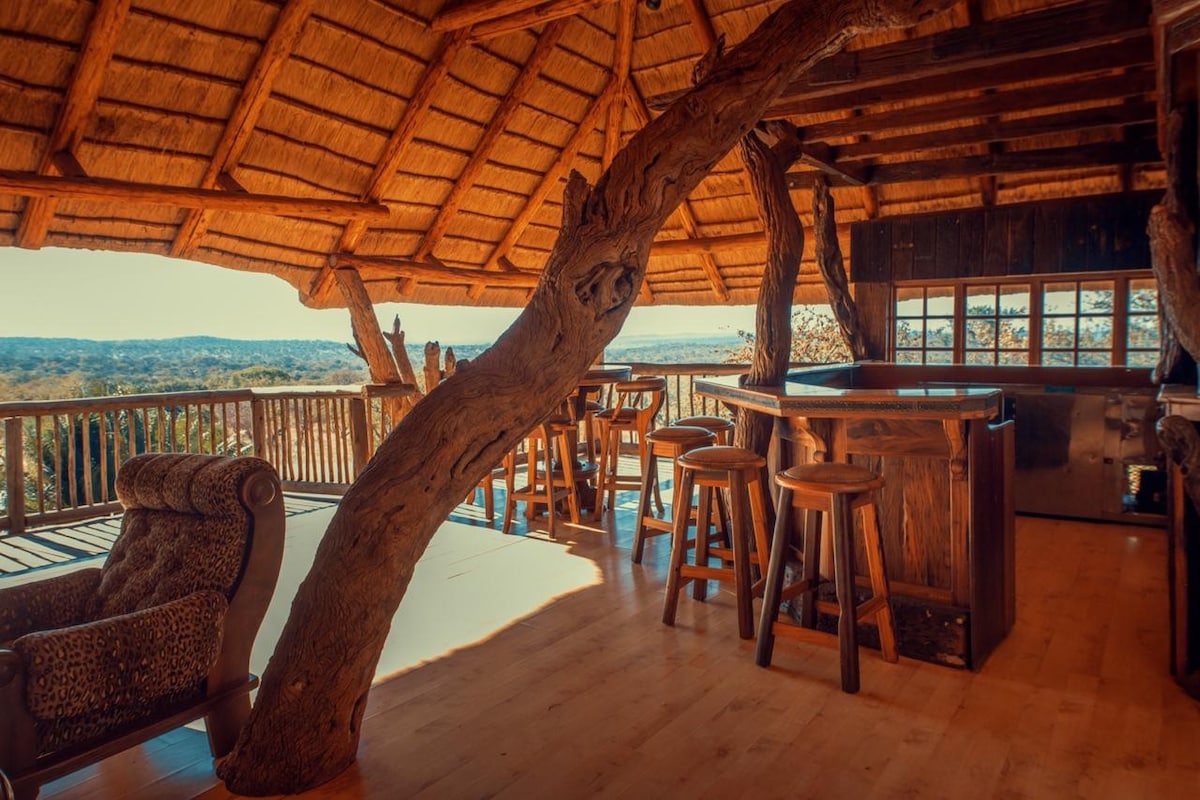 An open-air space features a thatched roof supported by natural wooden beams. A rustic bar with high stools stands prominently, accompanied by a cozy armchair nearby. Large windows offer expansive views of the surrounding landscape, creating a bright and inviting atmosphere.