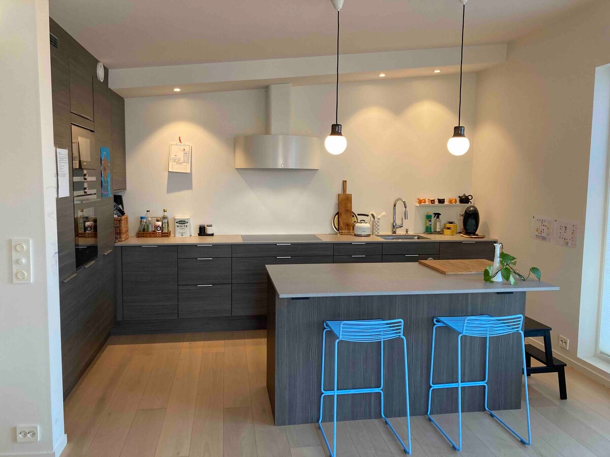 A modern kitchen features dark cabinetry and a spacious island equipped with two blue stools. Under-cabinet lighting illuminates the countertops, while pendant lights enhance the airy atmosphere. Various kitchen appliances and utensils are neatly arranged, and a plant adds a touch of greenery.