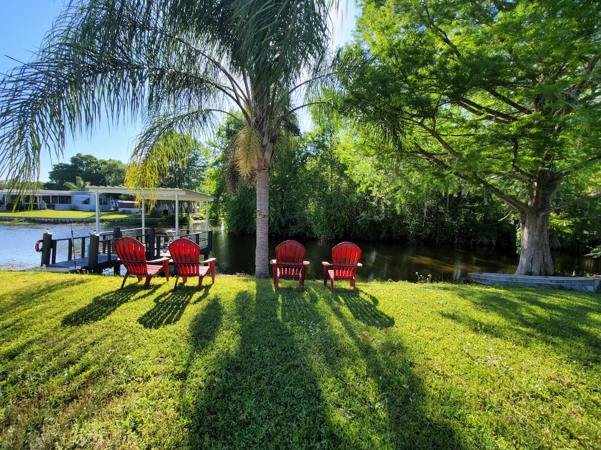 Four red Adirondack chairs are positioned on a grassy area, facing a calm body of water lined with lush trees. The sun casts long shadows, creating a serene and inviting outdoor space.