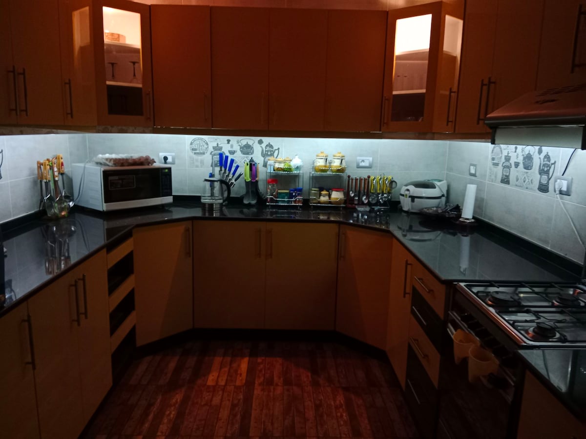 A well-equipped kitchen is shown with sleek cabinets and a dark countertop. Shelves display an array of kitchenware, including utensils and jars. Under-cabinet lighting provides illumination, creating a functional space for meal preparation. Appliances such as a microwave and stove are also visible.