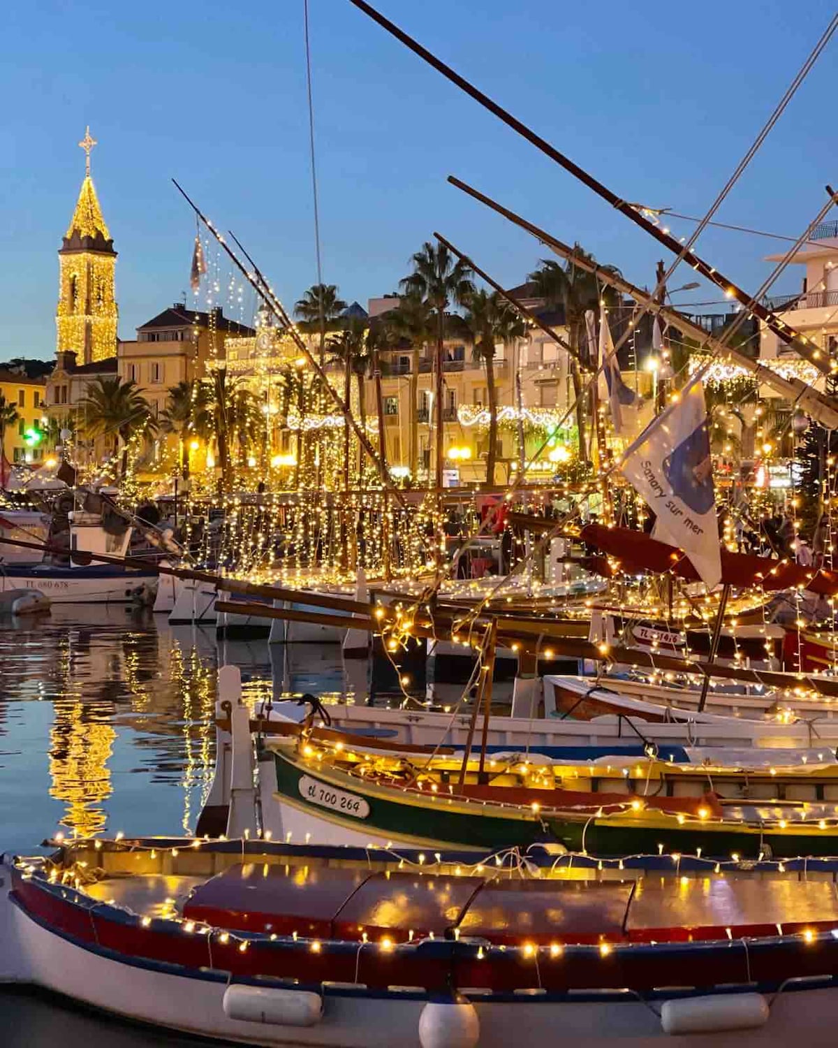 The harbor is adorned with boats decorated in twinkling lights, reflecting on the calm water. In the background, the illuminated buildings and a clock tower contribute to a festive atmosphere, surrounded by palm trees and warm evening lights.