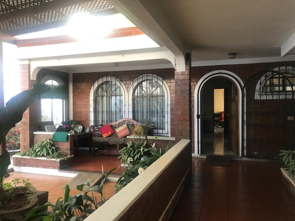 A welcoming entrance is visible, featuring arched windows and a covered porch adorned with vibrant cushions. Lush greenery and potted plants enhance the space, creating a serene setting. The layout leads toward an inviting living area, indicated by a glimpse of the interior beyond the entrance.