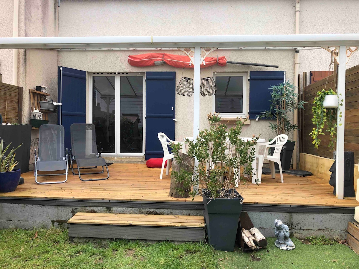 A wooden deck area is shown, featuring a seating arrangement with two lounge chairs and a small table. Potted plants are positioned around, and sliding glass doors lead to the interior. Blue shutters adorn the windows, and a red cover is draped overhead.