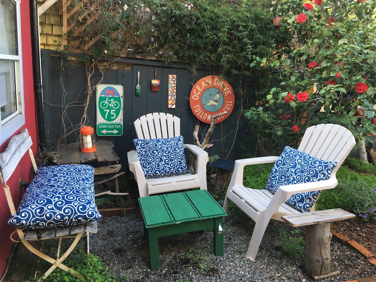 A quaint outdoor sitting area features two white chairs with blue patterned cushions and a green table. Surrounding greenery and blooming flowers enhance the space's charm. A bicycle route sign adds a local touch, while decorative wall features create a welcoming atmosphere.