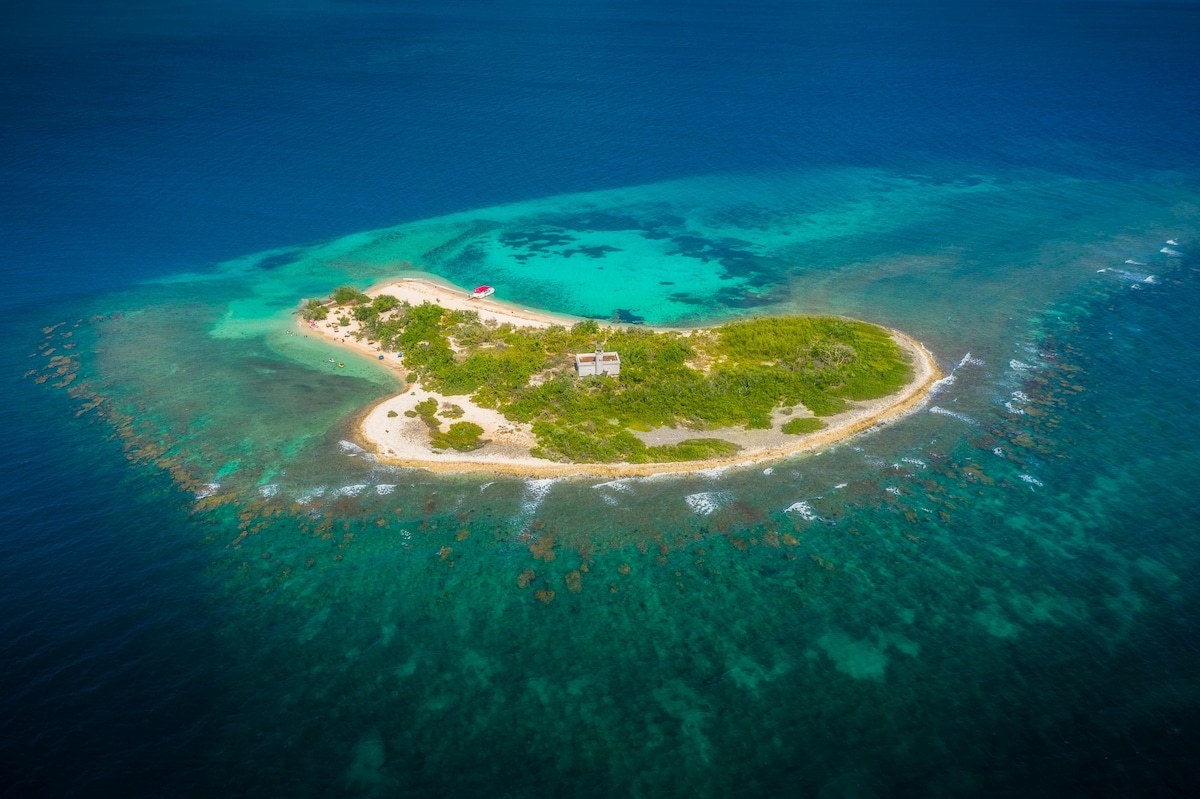 Aerial view of a remote island surrounded by vibrant blue waters. The island features a mix of sandy beaches and lush greenery, with coral reefs visible beneath the clear water. A small building is positioned amidst the natural landscape, offering a sense of seclusion and tranquility.