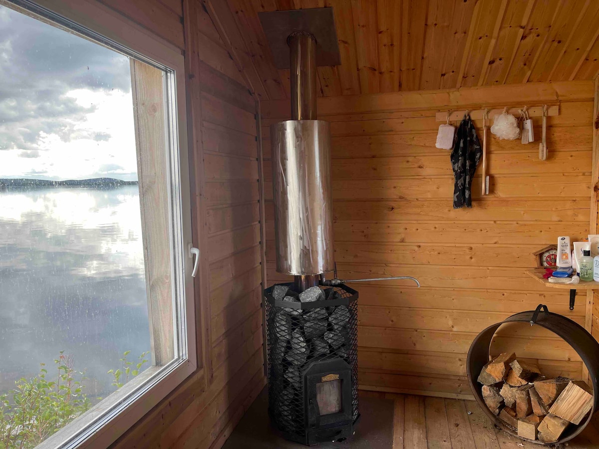 A wood-burning sauna stove is positioned against a wooden wall, with a window offering views of the nearby lake. A basket filled with firewood is located on the floor, while various sauna accessories hang neatly from hooks on the wall.