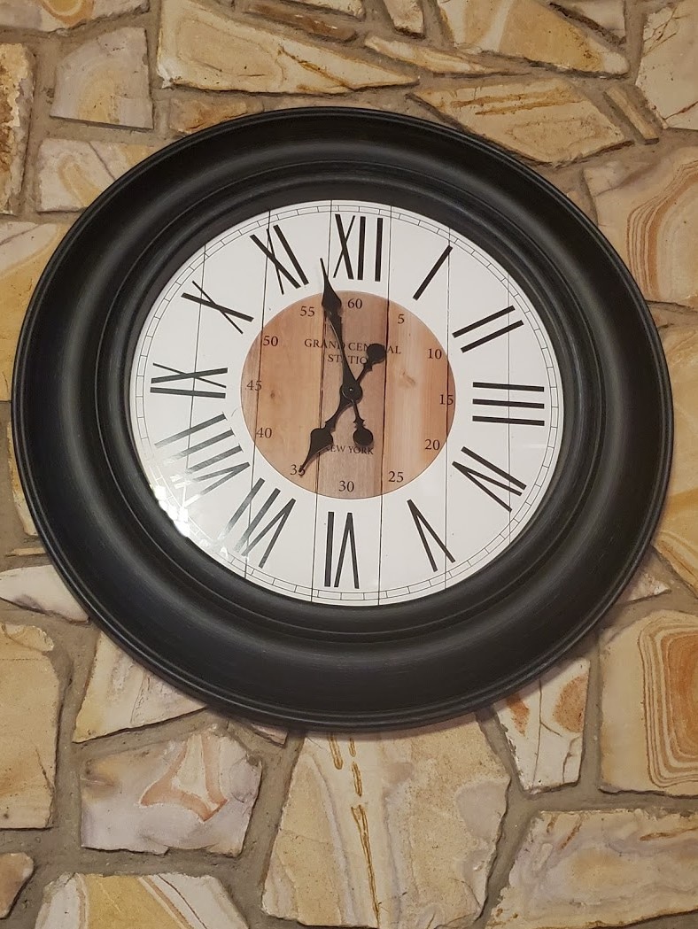 A round wall clock is mounted against a stone backdrop. The clock features a classic design with Roman numerals displayed on a white face and a wooden center. The black frame enhances its elegant appearance.