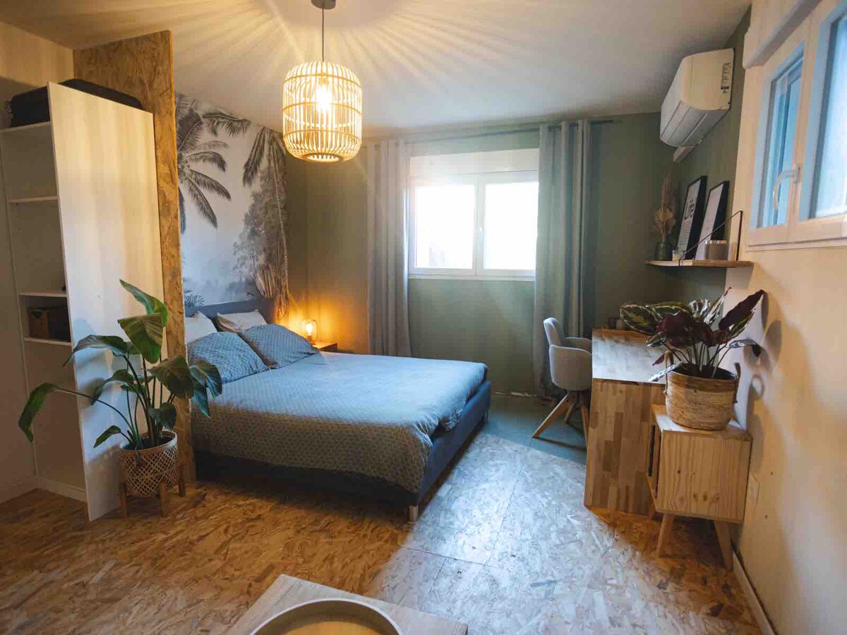 The room features a spacious bed with light bedding, complemented by a decorative wall mural of palm trees. A desk and chair are positioned near a window, with curtains gently framing the view. Potted plants add a touch of greenery to the decor.