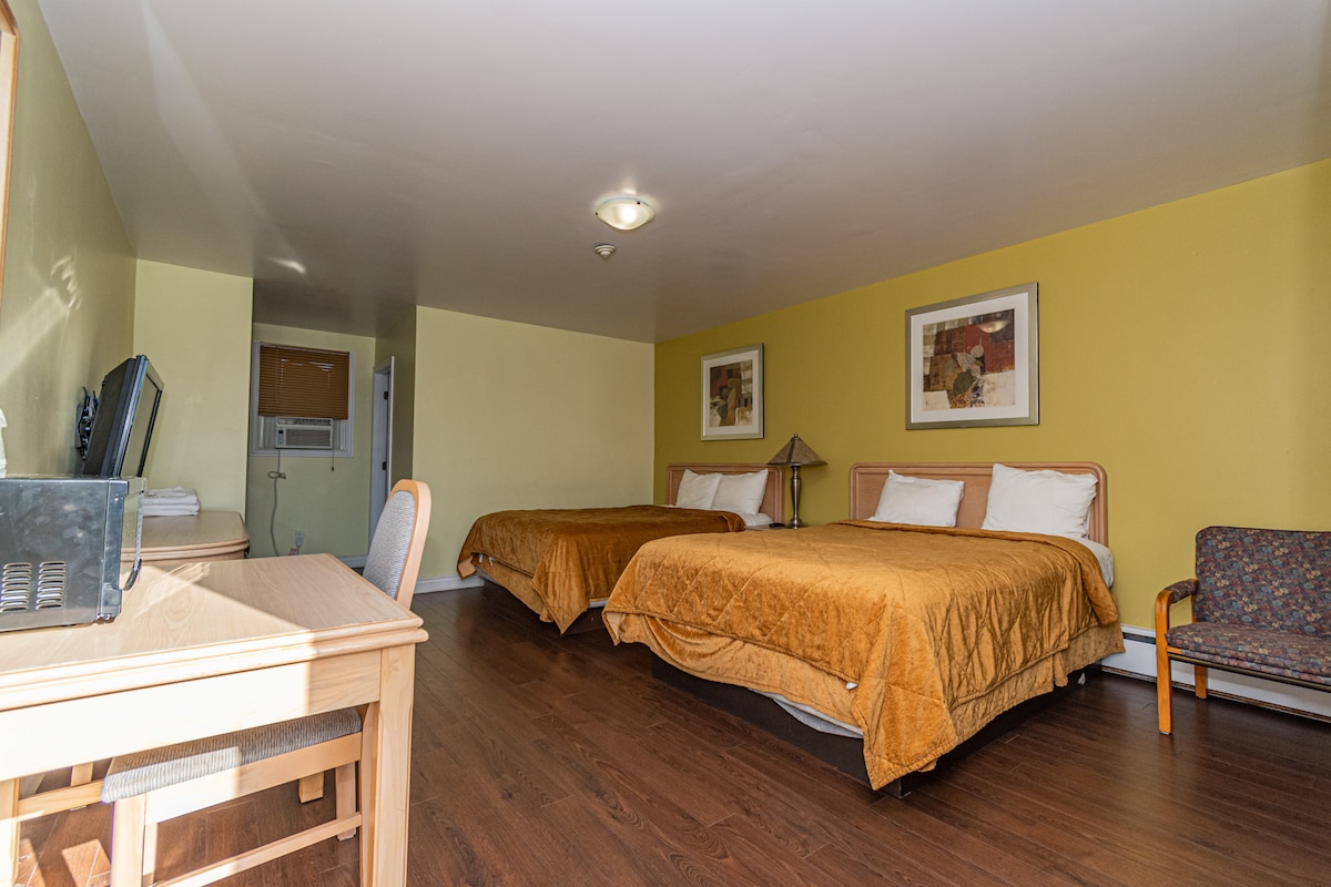 The family room features two queen-size beds with warm gold bedding. A wooden desk and chair are positioned beside a colorful, upholstered chair. The room includes a flat-screen TV and artwork on the walls, with light filtering in from a window across the room.