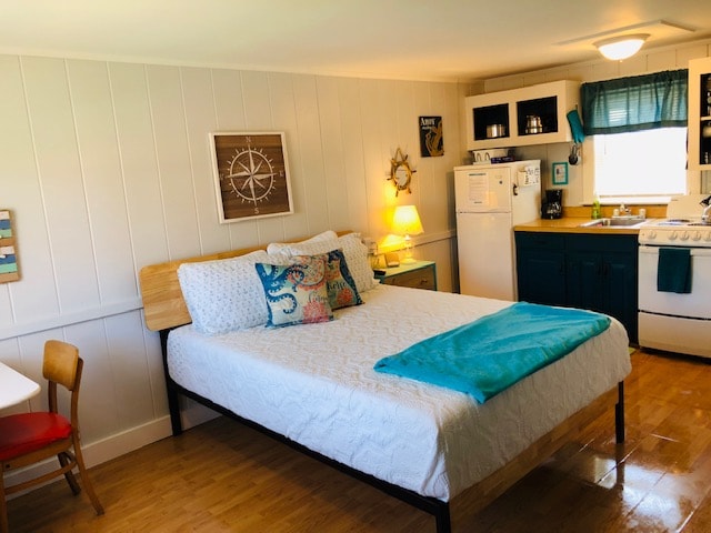 A functional studio layout features a queen bed with a light blue throw. A kitchenette with a stove, refrigerator, and coffee maker is visible, along with a dining table and a chair. Natural light floods the space through a window dressed with light curtains.