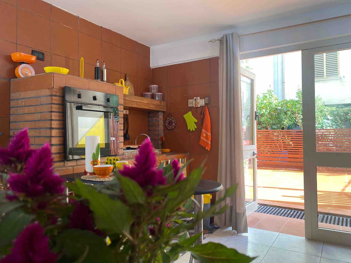 A well-equipped kitchen features a brick oven and vibrant colored utensils. Natural light fills the space through a large door leading to an outdoor area. A bouquet of purple flowers adds a touch of color to the foreground, creating a cheerful atmosphere.