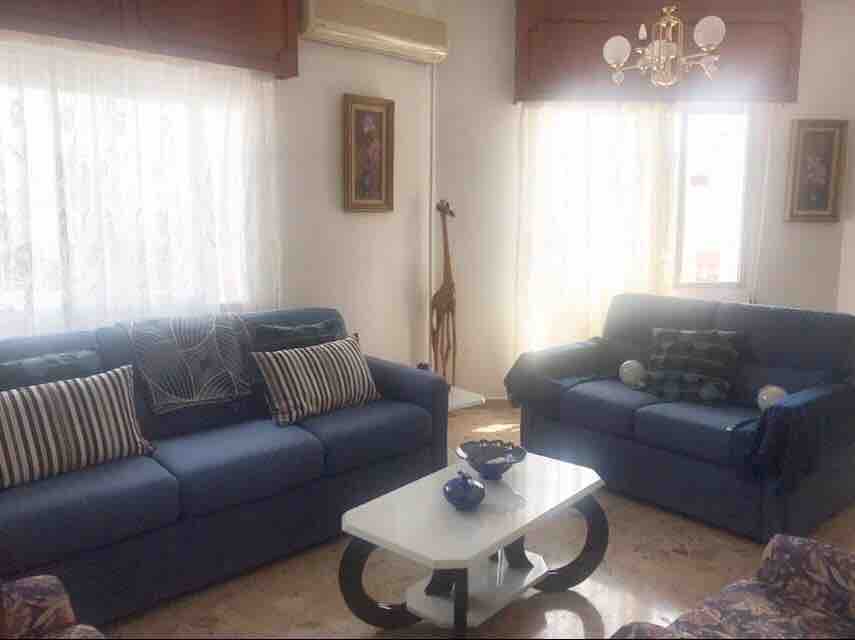 A bright living room features two blue sofas adorned with striped cushions, positioned around a central white coffee table. Natural light filters through sheer curtains, illuminating the space, while decorative wall art and a chandelier add a touch of elegance.