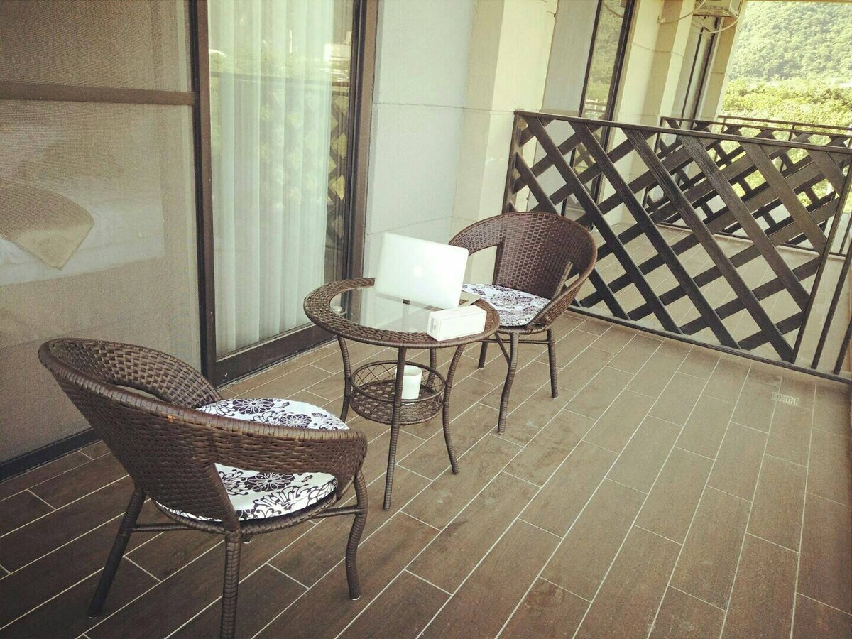 A private balcony features two wicker chairs with patterned cushions surrounding a small round table. Large floor-to-ceiling windows provide a view of the surroundings, while a railing adds safety and a touch of elegance to the space.