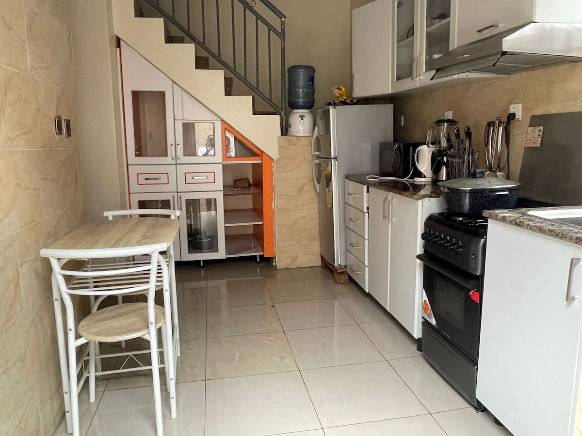An equipped kitchen is visible, featuring a stove, oven, and a countertop with essential appliances. A small dining table with two chairs is situated near the stairs, while storage cabinets and a water dispenser are present, providing convenience and functionality.