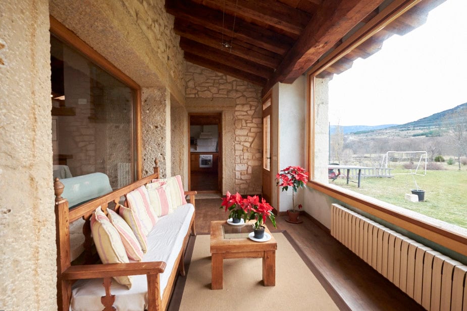 A sunlit sitting area features large windows offering views of the surrounding landscape. A wooden bench with cushion seating is paired with a low table, complemented by a potted flower arrangement. The rustic stone walls and wooden beams add charm to the inviting space.