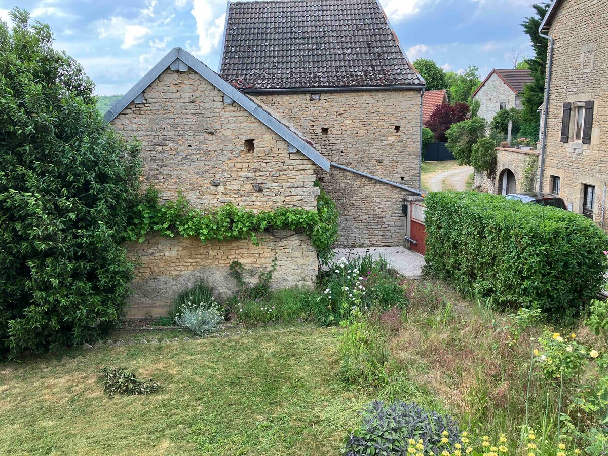A rustic stone building surrounded by green foliage and wildflowers is depicted. The structure features a pitched roof with shingles, and a pathway leads to a red door partially visible. The lawn area showcases a mix of grass and wild plants, indicating a natural landscape.
