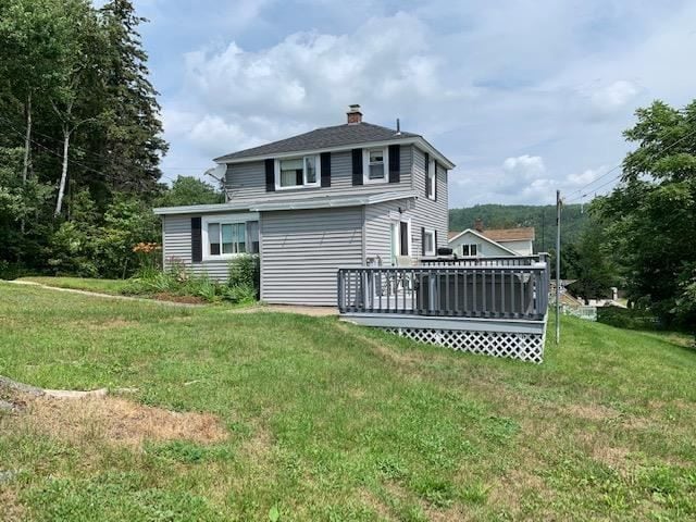 A two-story home is nestled in a green landscape, characterized by a lawn and an expansive deck at the back. The exterior is clad in light gray siding, with dark shutters adorning the windows. Surrounding trees complement the quiet, natural setting.