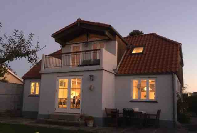 The house features a two-story exterior with a cozy balcony on the upper level. Large windows allow ample light to fill the interior, while the inviting patio area below includes outdoor seating. The warm glow from inside suggests a welcoming ambiance during twilight.