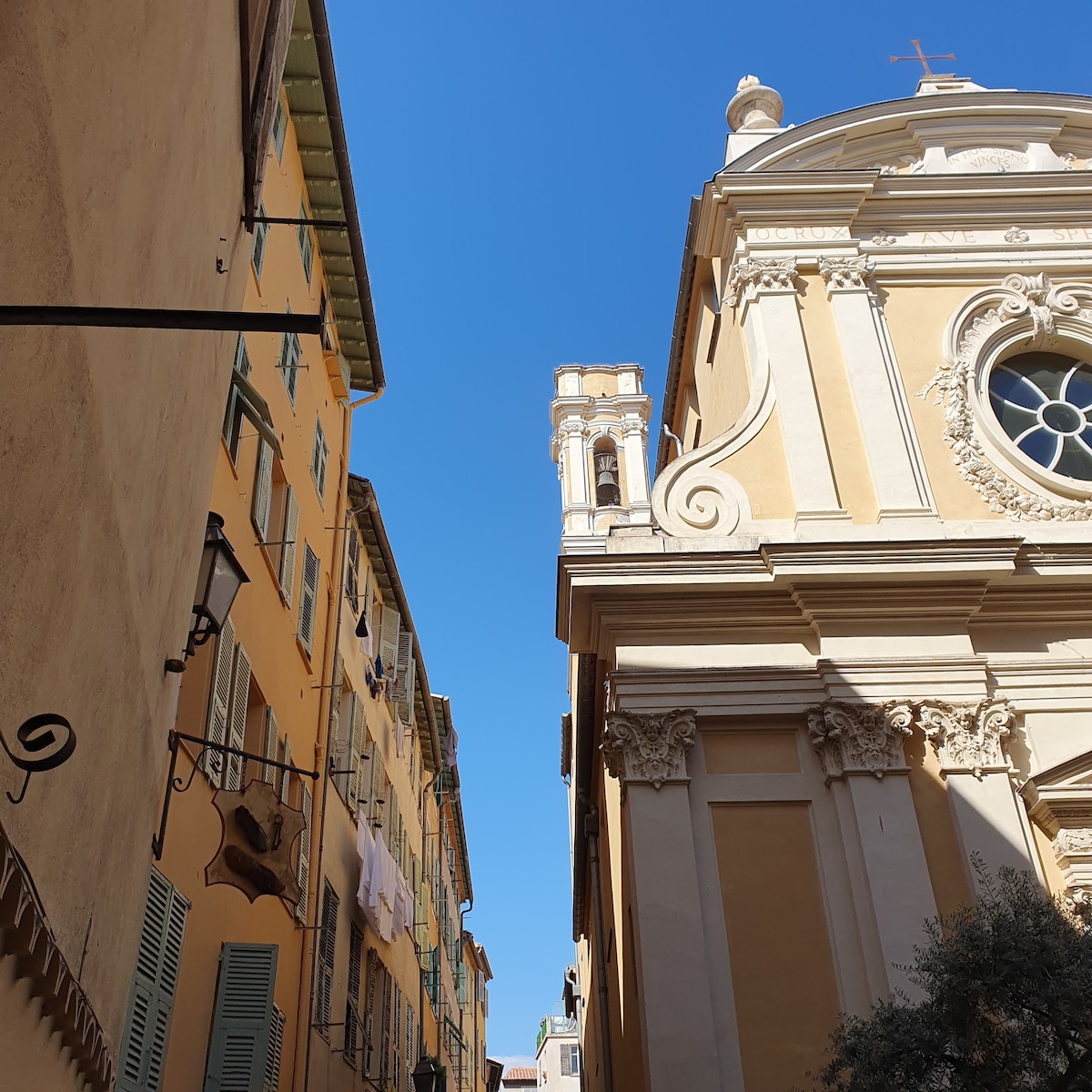 A narrow street is framed by tall, pastel-colored buildings. One building features a prominent church, showcasing intricate architectural details and a large round window. Clear blue skies create a bright backdrop, enhancing the charm of the local neighborhood.