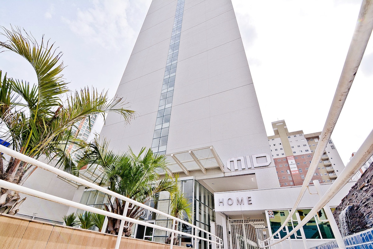 The modern building features a sleek architectural design with a prominent entrance displaying the word 'HOME.' Lush palm trees frame the entrance, contributing to a welcoming ambiance. The facade includes large glass windows that reflect the surrounding area.