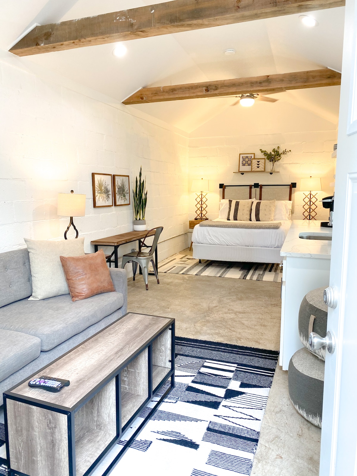 A studio apartment features a comfortable seating area with a gray sofa and a wooden coffee table. The sleeping area includes a spacious bed flanked by decorative lamps. Natural light enters through a window, illuminating a neutral color palette and wooden ceiling beams.