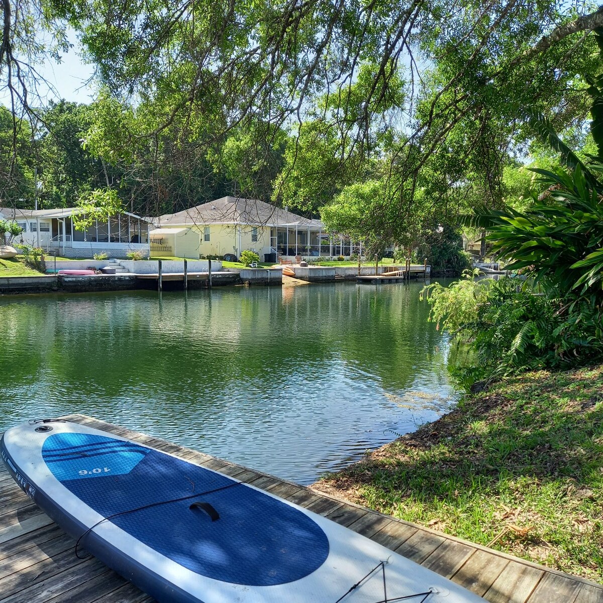 A paddleboard is positioned at the water's edge, with a calm canal reflecting surrounding greenery and a spacious home in the background. Lush trees provide shade, while the serene waters are complemented by hints of nearby kayaks and a tranquil, inviting atmosphere.