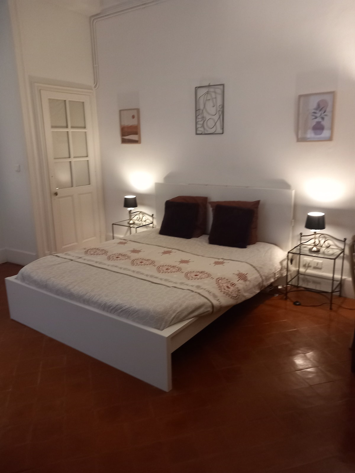 A well-lit bedroom features a spacious bed with a decorative coverlet and two pillows. Nightstands with lamps sit on either side, enhancing the ambience. Light-colored walls and warm-toned flooring contribute to the overall serene atmosphere.