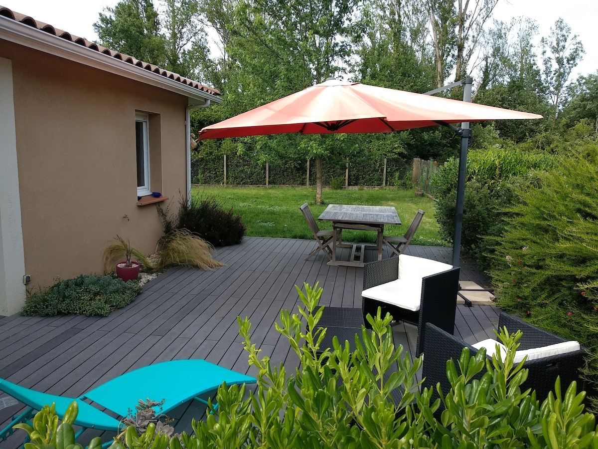 A patio area is set up with a large orange umbrella providing shade over a wooden table surrounded by chairs. A comfortable seating arrangement includes two black lounge chairs. Lush greenery and trees are visible in the background, creating a serene outdoor environment.