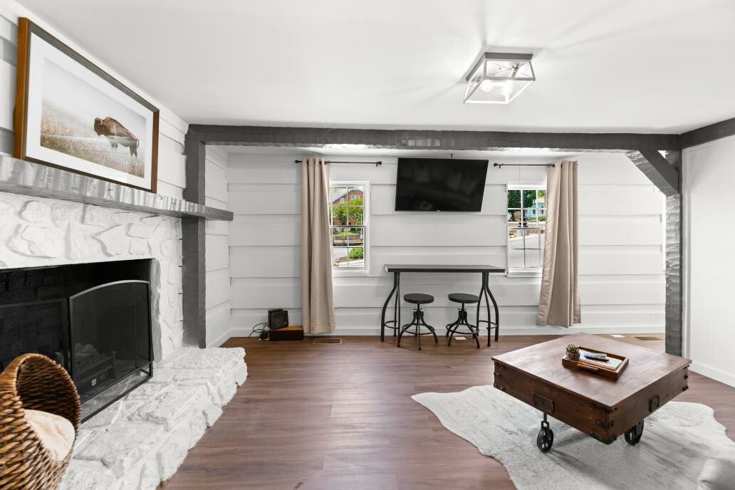 A cozy living area features a stone fireplace alongside a modern television mounted on the wall. Two large windows are dressed with light curtains, allowing natural light. A rustic coffee table sits on a light rug, enhancing the warm ambiance of the space.