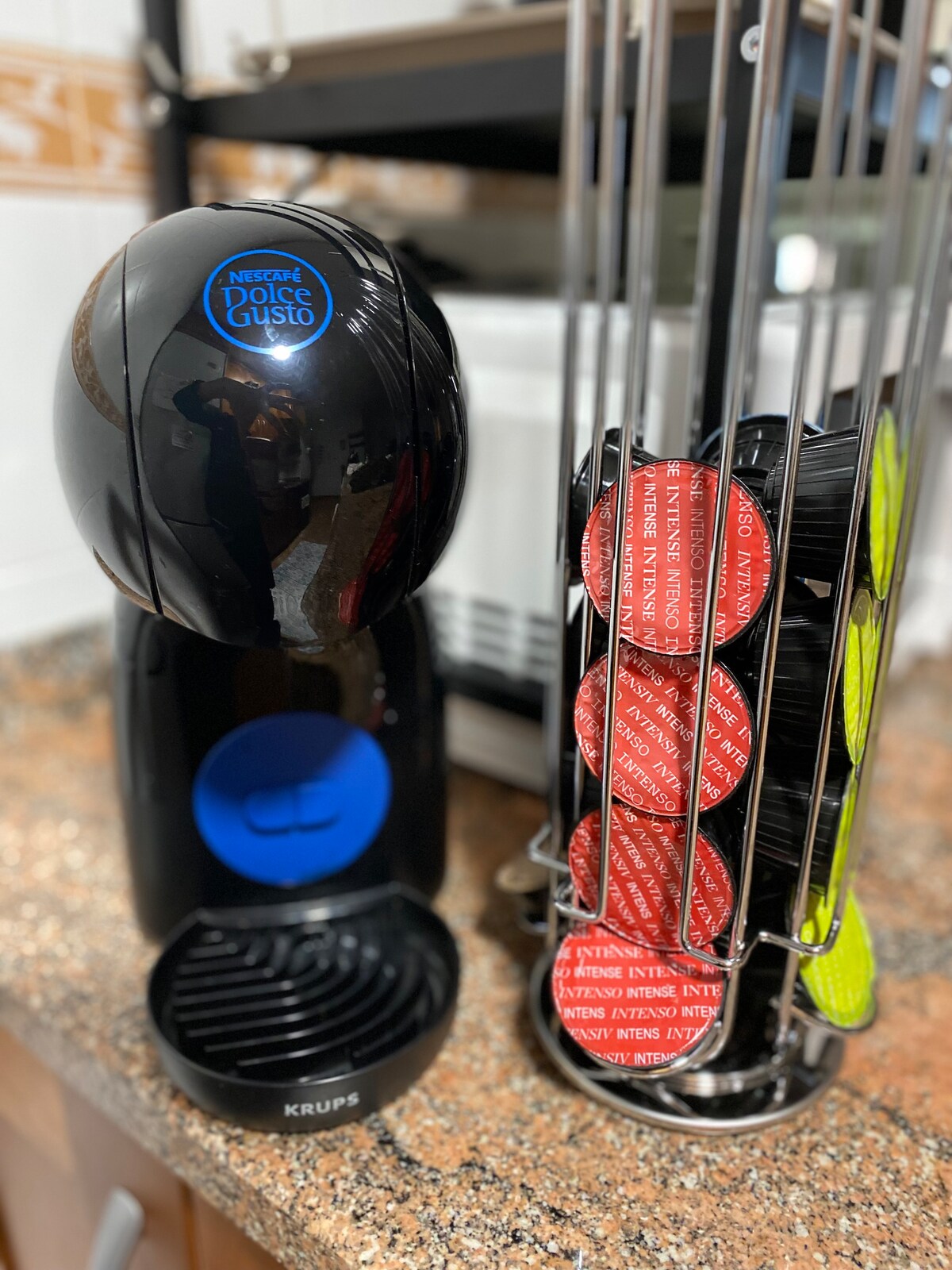 A coffee machine is positioned on a textured countertop, accompanied by a capsule holder featuring multiple coffee pods in black and red. The machine is sleek and modern, with a prominent blue accent and an arranged reservoir for used capsules.
