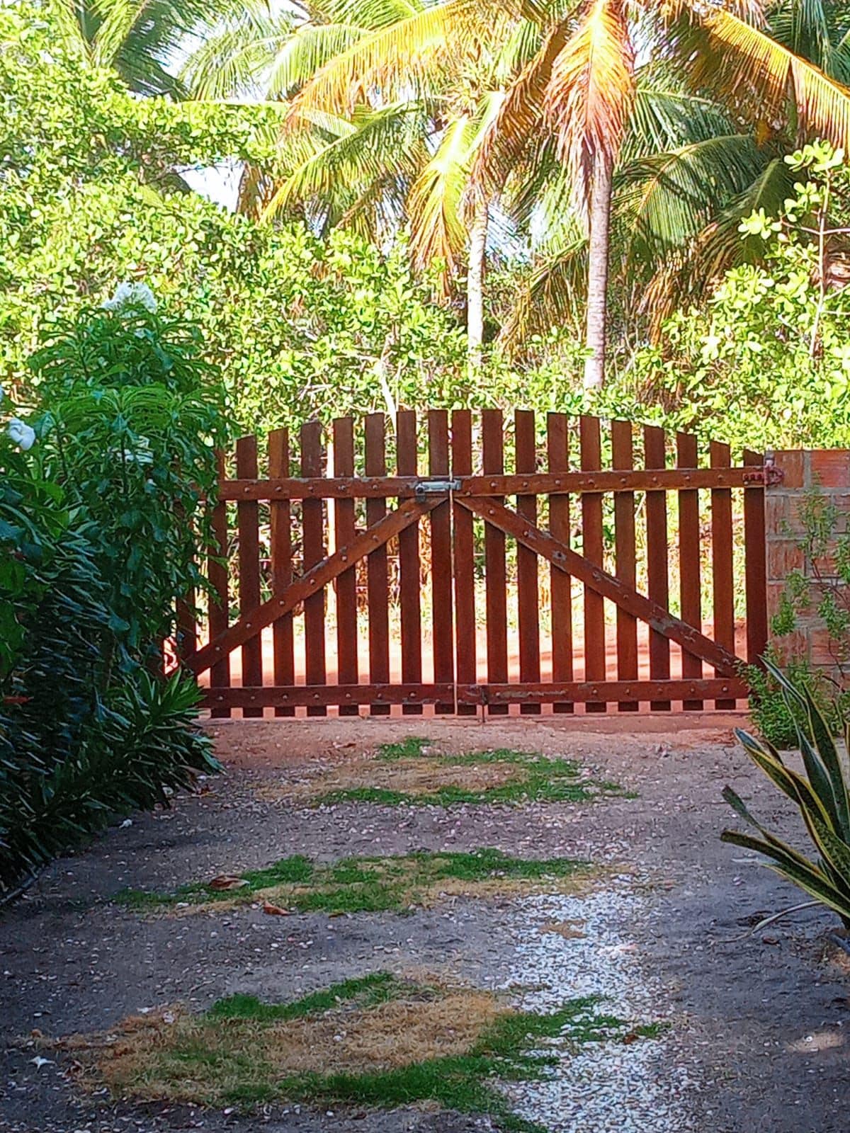 A wooden gate is set against a backdrop of lush greenery, including palm trees and other foliage. A gravel pathway leads up to the gate, with patches of grass visible along the ground. The bright sunlight enhances the natural colors of the surroundings.