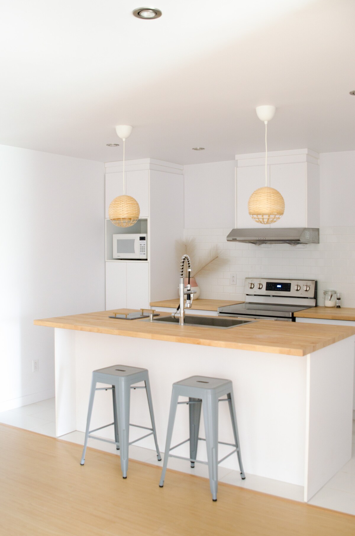 A modern kitchen features a light wooden countertop with a stainless-steel sink and stove. Two metal bar stools are placed at the counter. Pendant lights hang from the ceiling, providing illumination. The space is bright and airy, with white cabinetry and minimalist design elements.
