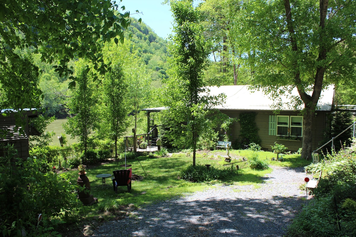 The cottage is framed by lush greenery, featuring a gravel pathway leading to a covered porch. The surrounding yard includes seating options and decorative elements nestled among the trees, creating a tranquil outdoor space with views of the nearby river and hills.