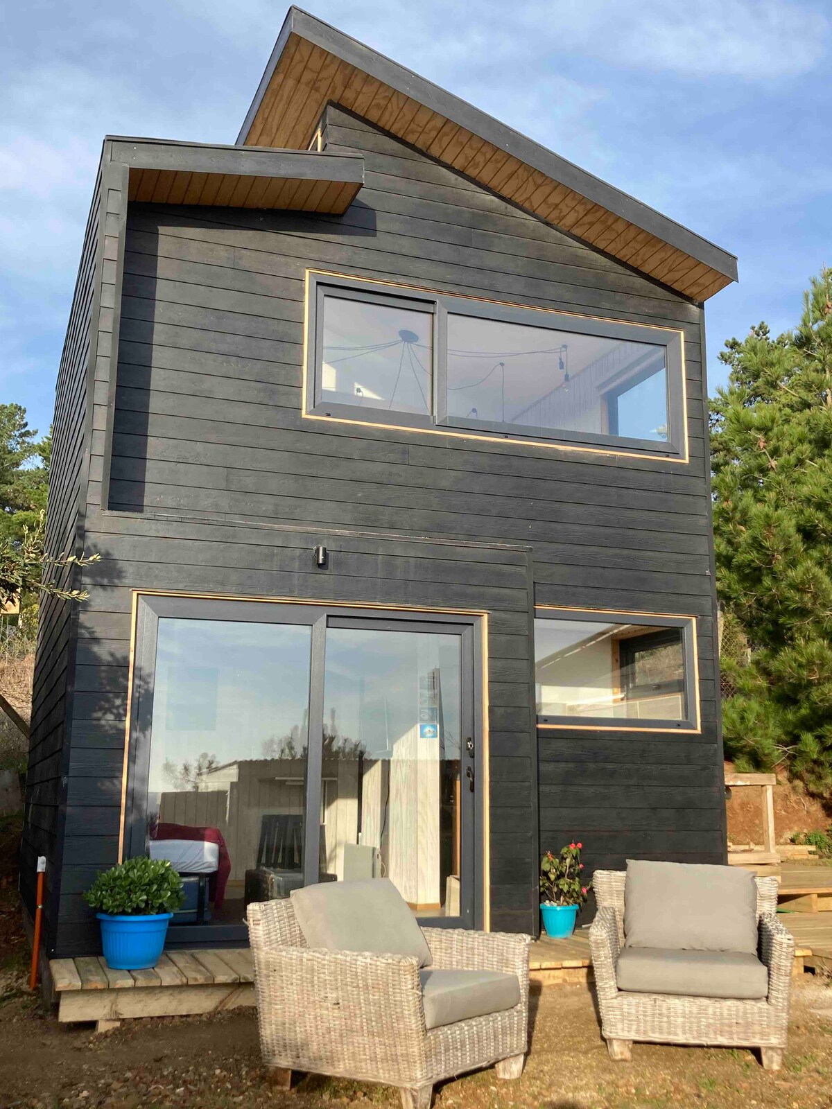 A modern two-story tiny house is depicted, featuring dark exterior wood cladding. Large glass doors provide an expansive view of the interior. Two comfortable wicker chairs are positioned outside, accompanied by a potted plant, enhancing the outdoor seating area.