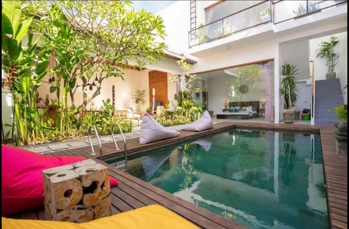 A serene outdoor area features a rectangular pool surrounded by wooden decking. Colorful bean bags are positioned by the water's edge, complemented by tropical plants. A relaxation area is visible in the background, with natural light enhancing the inviting atmosphere.