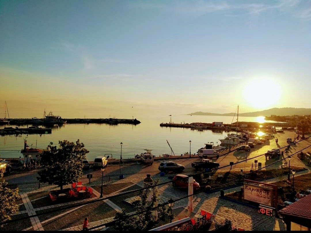 A serene waterfront scene is captured, showing calm waters reflecting the hues of sunrise. Boats are docked at the pier, while a tree-lined promenade invites leisurely strolls. The shoreline is lined with quaint establishments, enhancing the vibrant atmosphere of the harbor.