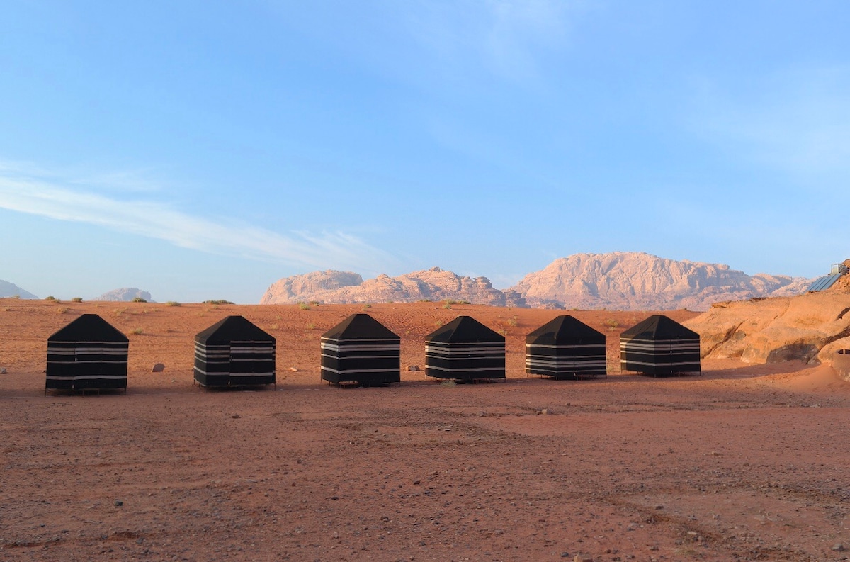 A series of traditional black tents is arranged in a line, set against a backdrop of arid desert landscape and distant mountains. The tents are characterized by their distinctive design, blending with the natural environment under a clear blue sky.